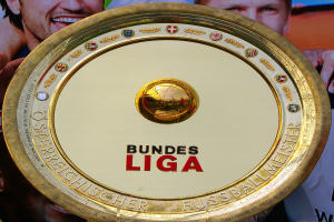 Ein Mann mit einem goldenen Schild mit der Aufschrift 'Bundesliga', im Hintergrund eine Fahne mit drei Personen.