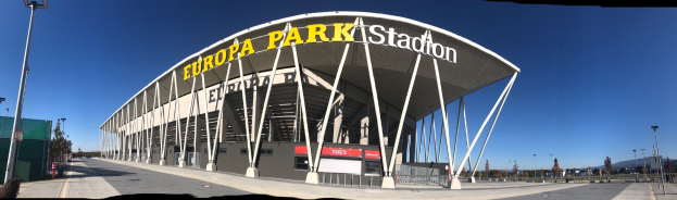 Großes Stadiongebäude mit Text, umgeben von Pfählen, Lichtern, Bäumen und einem Zaun unter einem sichtbaren Himmel.