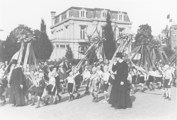 Schwarzes und weißes Bild einer Gruppe von Menschen, die eine Straße entlanggehen und Fahnen halten, mit Hüten, Bäumen, Gebäuden und einem Maibaum im Hintergrund.
