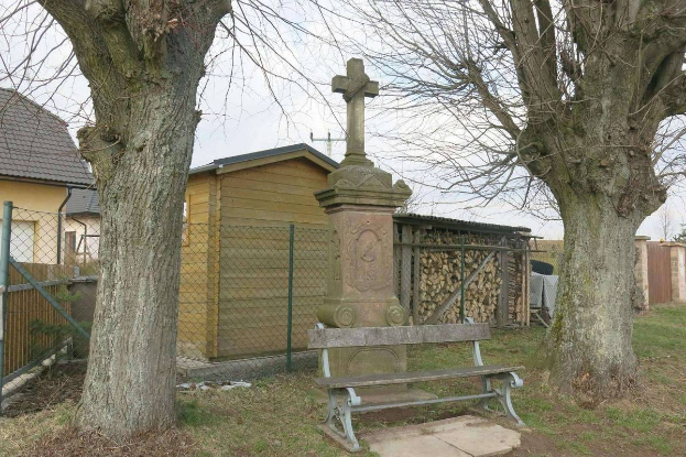 Eine Bank in einem grünen Feld neben einem Baum, mit einem Kreuz auf einem Pfeiler im Vordergrund, Häusern, einem Zaun und einem klaren blauen Himmel im Hintergrund, markiert das Grab eines Mannes, der bei einem Autounfall in St. Louis, Missouri, ums Leben kam.