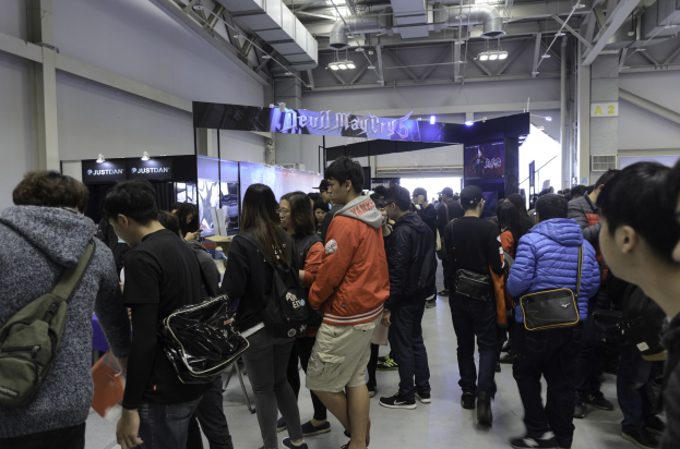 Eine Gruppe von Menschen steht vor einer größeren Menge an einem Ort, der wie eine Gaming-Messe aussieht, mit Schildern, Lichtern und Rohren im Hintergrund.