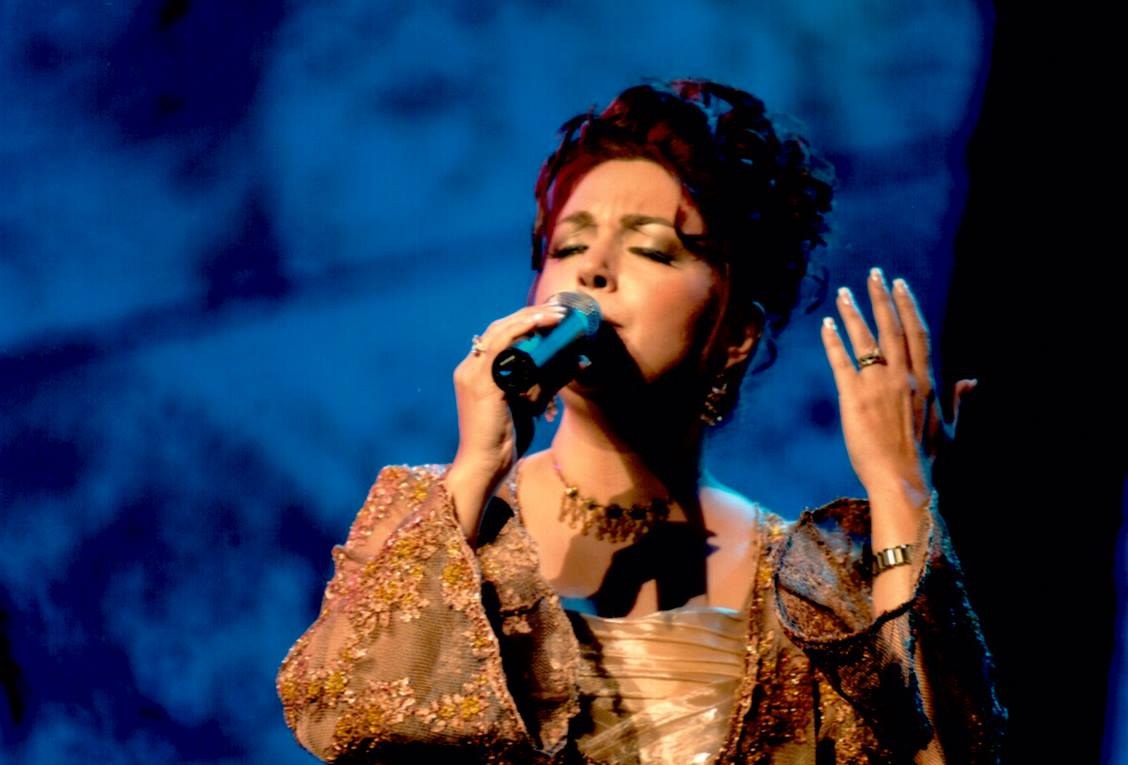 Frau in goldenem Kleid, singend ins Mikrofon auf der ESC-Bühne mit blau-schwarzem Hintergrund.