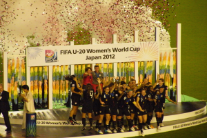 Gruppe von Frauenfußballspielerinnen in schwarzen Uniformen, die auf einer Bühne umgeben von einer Menge feiern, mit einer Tafel und dekorativen Gegenständen im Hintergrund.