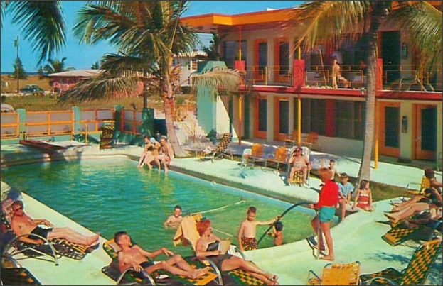 Ein Schwimmbad mit Menschen auf Stühlen im Vordergrund und Gebäuden, Bäumen und Autos im Hintergrund.