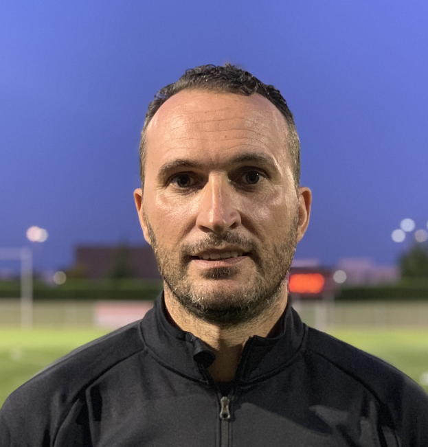 Fußballtrainer in schwarzer Jacke auf einem Fußballfeld mit unscharfem Hintergrund.