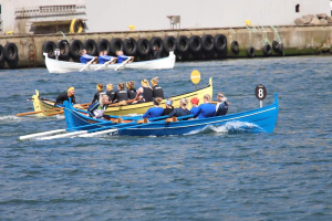 Eine Gruppe von Menschen in einem blauen und gelben Boot auf dem Wasser, die Paddel halten, mit einer Wand aus Reifen und einem Gebäude im Hintergrund, was auf eine Regatta hindeutet.