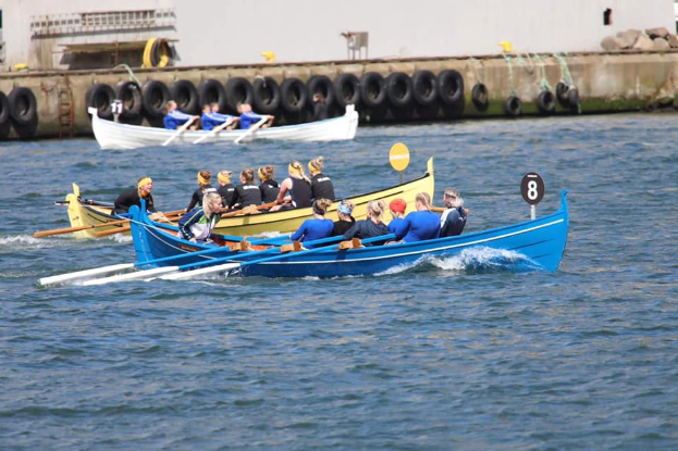 Eine Gruppe von Menschen in einem blauen und gelben Boot auf dem Wasser, die Paddel halten, mit einer Wand aus Reifen und einem Gebäude im Hintergrund, was auf eine Regatta hindeutet.