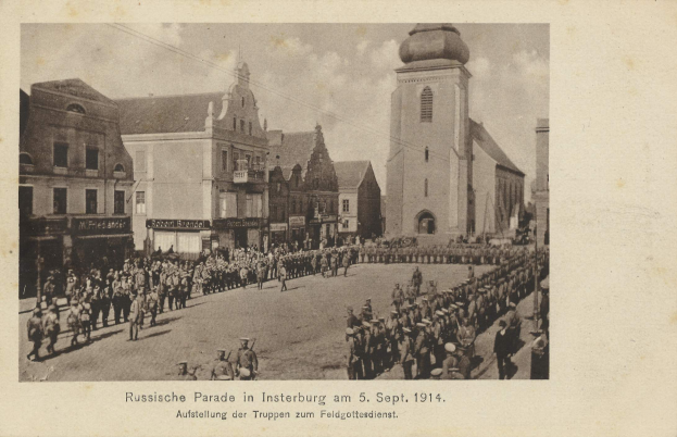 Ein Schwarz-Weiß-Foto eines Umzugs in Insterburg am 5. September 1914 mit vielen Menschen, Gebäuden im Hintergrund und Text unten.