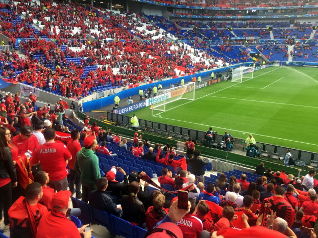 Eine große Menschenmenge in einem Stadion schaut einem Fußballspiel zu, mit Menschen auf Stühlen und Stehplätzen, Fahnen, einem Torpfosten und Deckenlichtern.