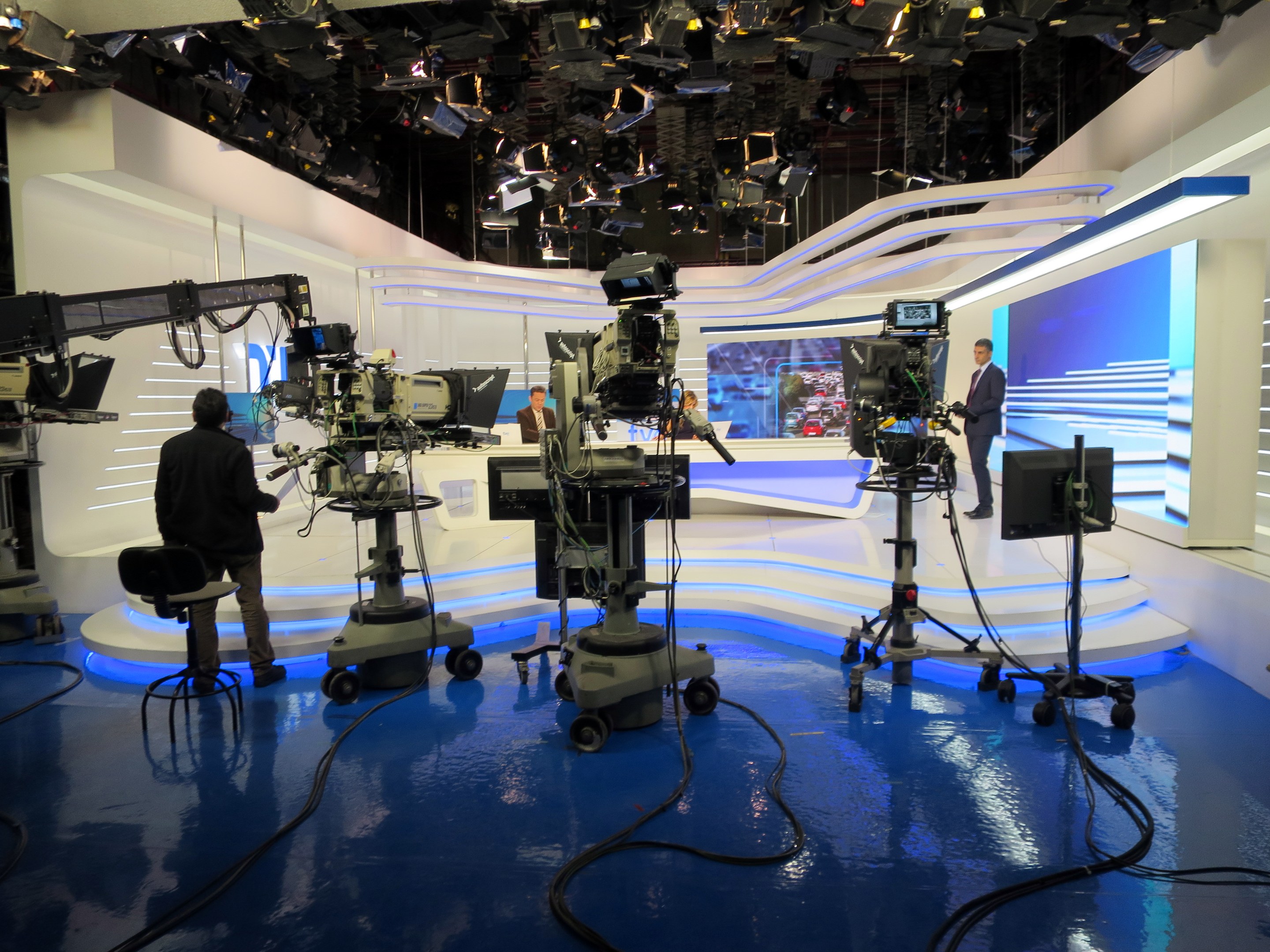 Ein TV-Studio mit mehreren Kameras auf dem Boden, einer sitzenden Person im Vordergrund und einigen stehenden Personen im Hintergrund, mit an der Decke montierten Lichtern für eine Live-Broadcast-Aufnahme.