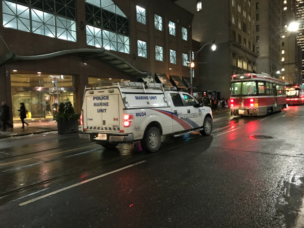 Ein Polizei-Lkw parkt nachts am Straßenrand, mit Fahrzeugen auf der Straße, Fußgängern auf dem Gehweg, Topfpflanzen, Straßenlaternen, Gebäuden mit Fenstern und einem bewölkten Himmel.