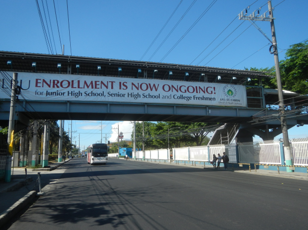 Ein Bus fährt unter einer Brücke mit einem "Jetzt Anmelden"-Schild, während Fußgänger darunter gehen und Strommasten die Straße säumen, mit Bäumen und einem klaren Himmel im Hintergrund.