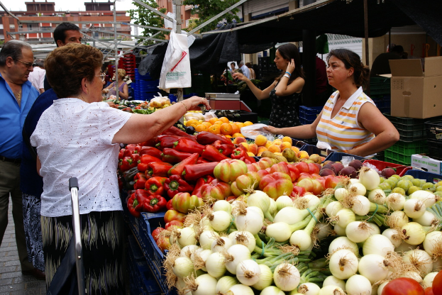 Eine Gruppe von Menschen, die sich draußen in der Nähe von Gebäuden und Bäumen aufhalten, mit Gemüse in Plastikbehältern und einer Kartonbox auf dem Boden.