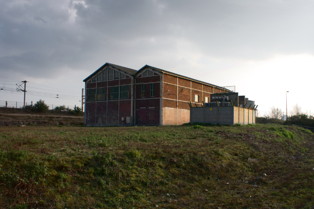 Eine Szene mit Schuppen, Transformatoren, Strommasten mit Kabeln, Zäunen, einem Straßenmast mit Straßenlaterne, Bäumen und einem bewölkten Himmel.