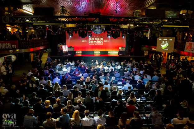 Eine große Menschenmenge sitzt auf Stühlen vor einer Bühne mit Bildschirmen und Bannern, die von Scheinwerfern beleuchtet wird und von Geländern umgeben ist, und erwartet gespannt den Beginn eines Online-Marketing-Rockstars-Events, mit einem Wasserzeichen in der linken unteren Ecke.