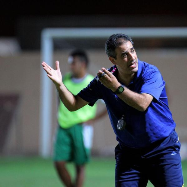 Ein Mann in einem blauen T-Shirt und Hose steht auf einem Fußballfeld und hält ein Mikrofon in der Hand, während er gestikuliert. Im Hintergrund sind eine Person in einem grünen Kleid, ein Tor und eine Wand zu sehen.