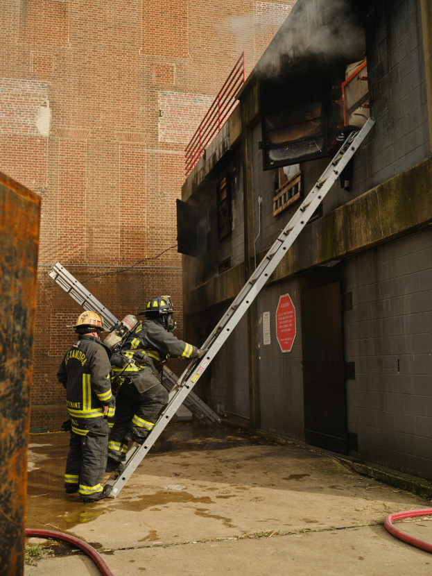 Feuerwehrleute mit Helmen klettern an einer Leiter zu einem rauchgefüllten Gebäude hoch, mit einem Metallobjekt links und sichtbaren Rohren am Boden.