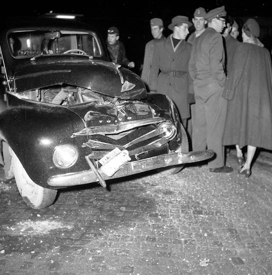 Schwarz-weißes Bild eines schwer beschädigten Autos am Straßenrand mit eingedellter Motorhaube und zersplitterten Fenstern, umgeben von Menschen, die die Szene begutachten.