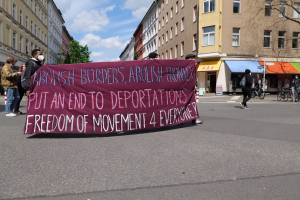 Eine Gruppe von Menschen marschiert mit einer Plakate, auf der "Abolish Borders, Abolish Frontiers, Put an End to Deportations, Freedom of Movement 4 Everyone" steht, durch eine Straße, mit Gebäuden, Bäumen und Fahrrädern an der Straße und einer bewölkten Himmelüber.