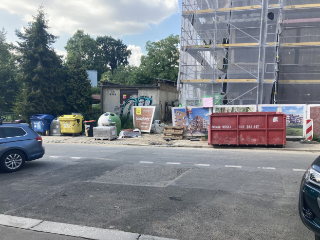 Zwei Autos neben einem Gebäude in Bau neben der Straße geparkt, mit verstreuten Mülltonnen, Brettern und anderen Gegenständen auf der Straße, Bäume im Hintergrund und ein bewölkter Himmel.