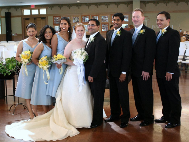 Eine Frau in einem weißen Kleid mit einem Strauß steht in der Mitte, lächelt und ist von Frauen in blauen Kleidern und Männern in schwarzen Anzügen umgeben, die auf einem Boden mit verschiedenen Gegenständen und Dekorationen im Hintergrund stehen.