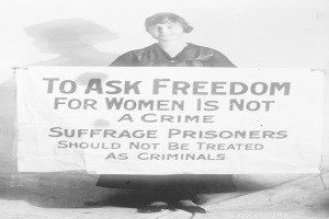 Schwarz-weiß-Fotografie einer Frau, die ein Banner hält, auf dem steht: "Für die Freiheit der Frauen zu kämpfen ist kein Verbrechen, Suffragetten sollten nicht wie Kriminelle behandelt werden", während sie lächelt.