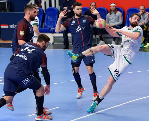 Eine Gruppe von Männern, die Handball auf einem Platz spielen, während Zuschauer von Stühlen, Geländern und einer Wand im Hintergrund zuschauen.