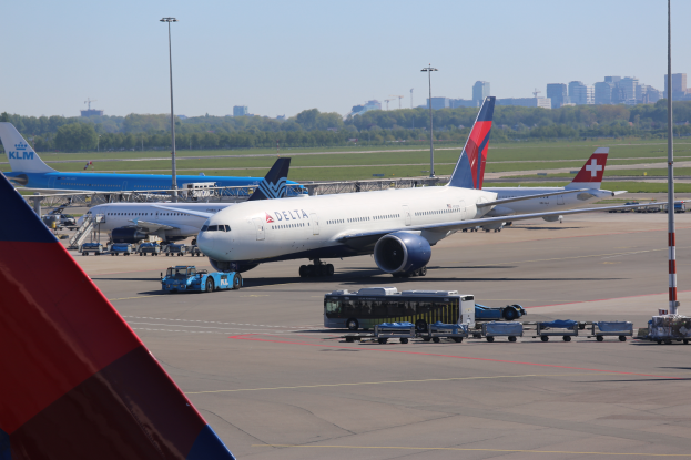 Ein Boeing 777-300ER der Delta Air Lines steht am Frankfurter Flughafen, umgeben von Fahrzeugen und Straßenlaternen, sowie Gebäuden, Bäumen und einem klaren blauen Himmel im Hintergrund.