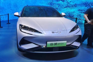 Ein weißes Auto auf einer Automesse mit einer Person rechts daneben, die ein Objekt hält, ein Bildschirm im Hintergrund und Absperrpoller mit Seilen links.