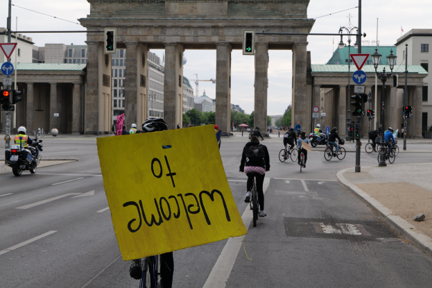 Eine Gruppe von Radfahrern fährt eine Straße vor dem Brandenburger Tor in Berlin, Deutschland, entlang, wobei einer ein gelbes Schild hält; die Szene umfasst Laternenpfähle, Verkehrsampeln, Gebäude, Bäume und einen klaren Himmel.