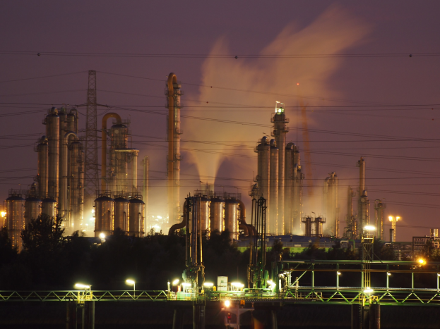 Ölraffinerie bei Nacht mit aufsteigendem Rauch, beleuchtet von nahen Lichtern, vor einem sichtbaren Himmel, mit einem Wasserzeichen.