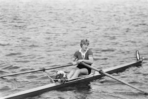 Eine Frau in einem weißen Hemd und blauen Shorts rudert ein Einerboot mit einem weißen Rumpf und blauer Segel auf einem sonnigen Gewässer, wobei sie zwei Paddel verwendet und ihr Haar zu einem Pferdeschwanz gebunden hat.