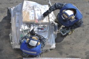Zwei Männer in blauen Uniformen und Helmen arbeiten an einem großen Drohnenmodell, jeder hält Werkzeuge in der Hand, mit einer Plastikabdeckung auf dem Boden.