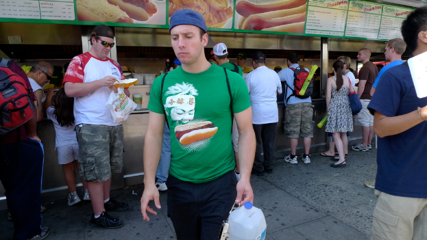 Ein Mann in einem grünen T-Shirt, schwarzen Hosen und einer blauen Mütze geht durch einen belebten Imbissbereich und hält eine Flasche in der Hand.
