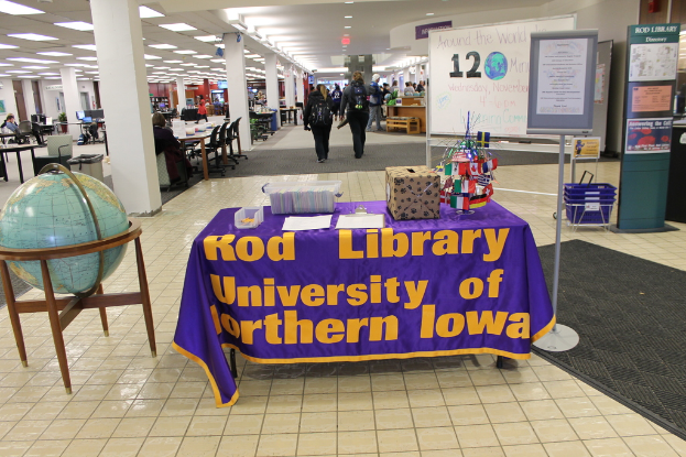 Eine Ausstellung mit einem Tisch, auf dem verschiedene Landesflaggen, eine Box, Papiere und ein Banner sowie links daneben eine Weltkugel und rechts daneben eine Anschlagtafel mit einigen Menschen in der Nähe und im Hintergrund mehrere Tische und Stühle zu sehen sind.