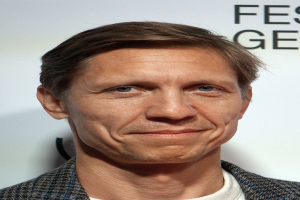 Ein Mann im Anzug und Krawatte lächelt in die Kamera, im Hintergrund eine Tafel mit der Aufschrift "Eddie Redmayne Net Worth"