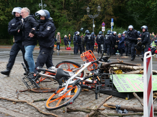 Eine Gruppe von Polizisten in schwarzen Uniformen und Helmen steht um einen Haufen Fahrräder auf der Straße, einige tragen Taschen, mit Laternenmasten, Schildern, Bäumen und einem klaren blauen Himmel im Hintergrund.