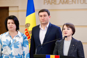 Zwei Frauen und ein Mann stehen an einem Podium vor einer ukrainischen Flagge, wobei der Mann ein schwarzes Sakko und ein weißes Hemd trägt und die Frau rechts ein aschfarbenes Kleid trägt und über die Ukraine-Krise diskutieren.