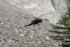 Ein Hubschrauber fliegt über eine felsige Bergseite, mit einem Baum auf der rechten Seite und einem Hügel im Hintergrund.