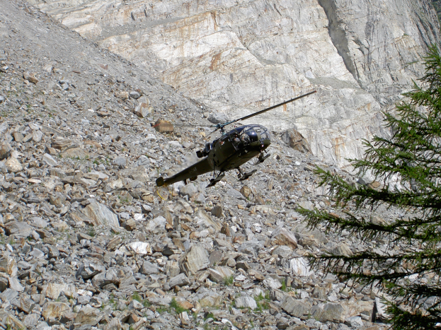 Ein Hubschrauber fliegt über eine felsige Bergseite, mit einem Baum auf der rechten Seite und einem Hügel im Hintergrund.
