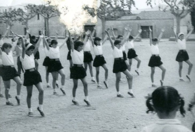 Schwarzes und weißes Bild von jungen Mädchen in Schuluniformen, die auf einer von Gebäuden und Bäumen gesäumten Straße tanzen, mit sichtbaren Personenschuhen am Boden.