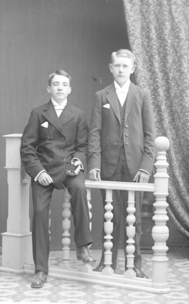 Zwei junge Männer in Anzügen stehen nebeneinander auf einem Balkon mit einem Geländer vorne und einem Vorhang im Hintergrund.