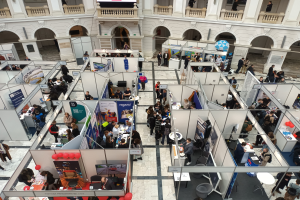 Große Besucherzahl auf einer internationalen Fachmesse mit Ständen, Bannern, Tischen, Stühlen und Luftballons in einem Raum mit Bogengängen, Säulen und einem Bildschirm im Hintergrund.