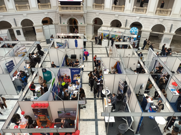 Große Besucherzahl auf einer internationalen Fachmesse mit Ständen, Bannern, Tischen, Stühlen und Luftballons in einem Raum mit Bogengängen, Säulen und einem Bildschirm im Hintergrund.