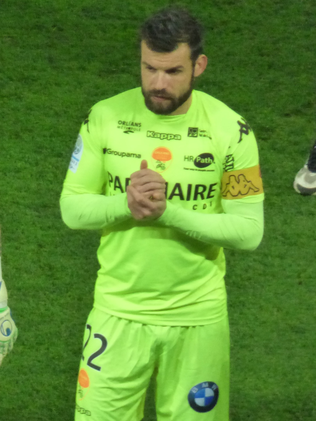 Torwart in gelbem Trikot klatscht auf dem Fußballfeld, trägt grünes Shirt und Shorts, mit dem Bein einer anderen Person links im Bild.
