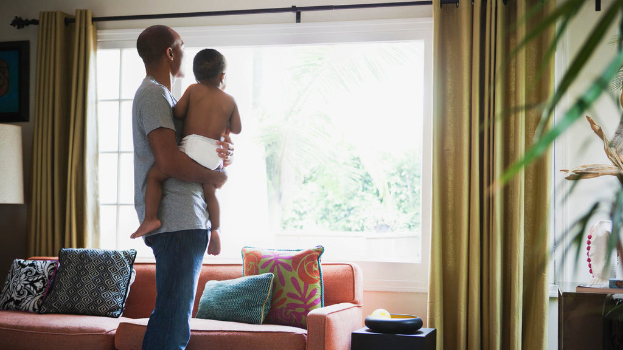Ein Mann trägt ein Kind vor einem Fenster mit Vorhängen, einer Couch und mehreren Kissen.