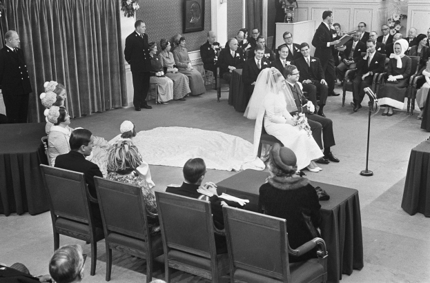 Schwarz-weiß-Foto einer Hochzeitszeremonie mit sitzenden und stehenden Menschen, einem Mann mit Mikrofon, einer Frau mit einem Bukett, einem Podium mit Mikrofon, Blumenschmuck, einem Vorhang, einem gerahmten Foto an der Wand und einer Tür im Hintergrund.