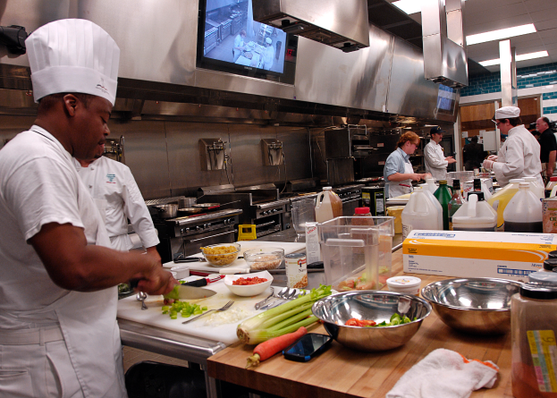 Eine Gruppe von Köchen in einer Küche, die Essen auf einem Schneidbrett zubereiten, mit einem Koch im Vordergrund, der eine weiße Schürze und eine Mütze trägt, ein Messer hält und von verschiedenen Utensilien, Schüsseln, Gemüse, einem Handy, Flaschen und anderen Gegenständen umgeben ist, während im Hintergrund ein Fernseher zu sehen ist.