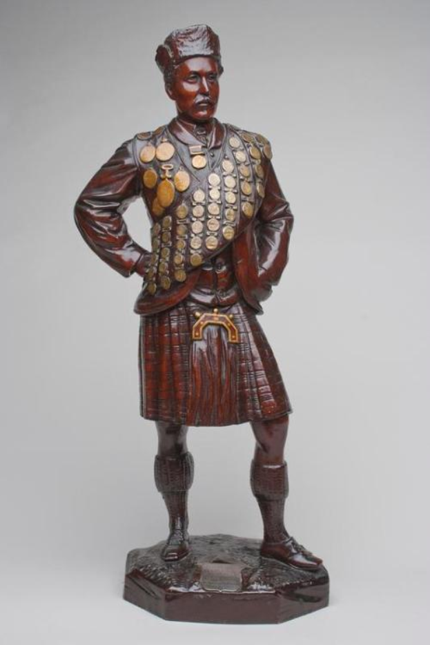 Eine hölzerne Skulptur eines Mannes in traditioneller schottischer Kleidung, einschließlich eines Tartan-Kilts, eines hochgeschlossenen Westen und eines Gürtels mit einer Schnalle, der selbstbewusst vor einem weißen Hintergrund steht.