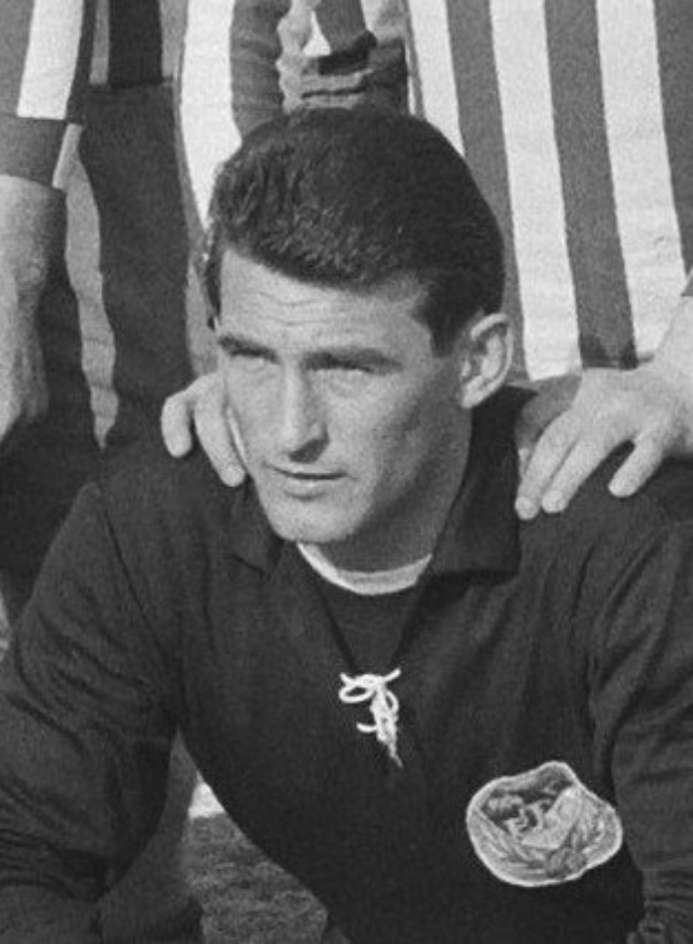 Schwarzes und weißes Foto eines Mannes, der auf einem grasbewachsenen Fussballfeld sitzend eine T-Shirt und eine Kette trägt, mit zwei Personen dahinter.